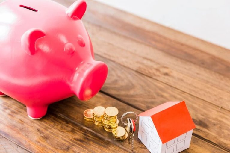 Monedas apiladas junto a alcancía de cerdo y figura de casa